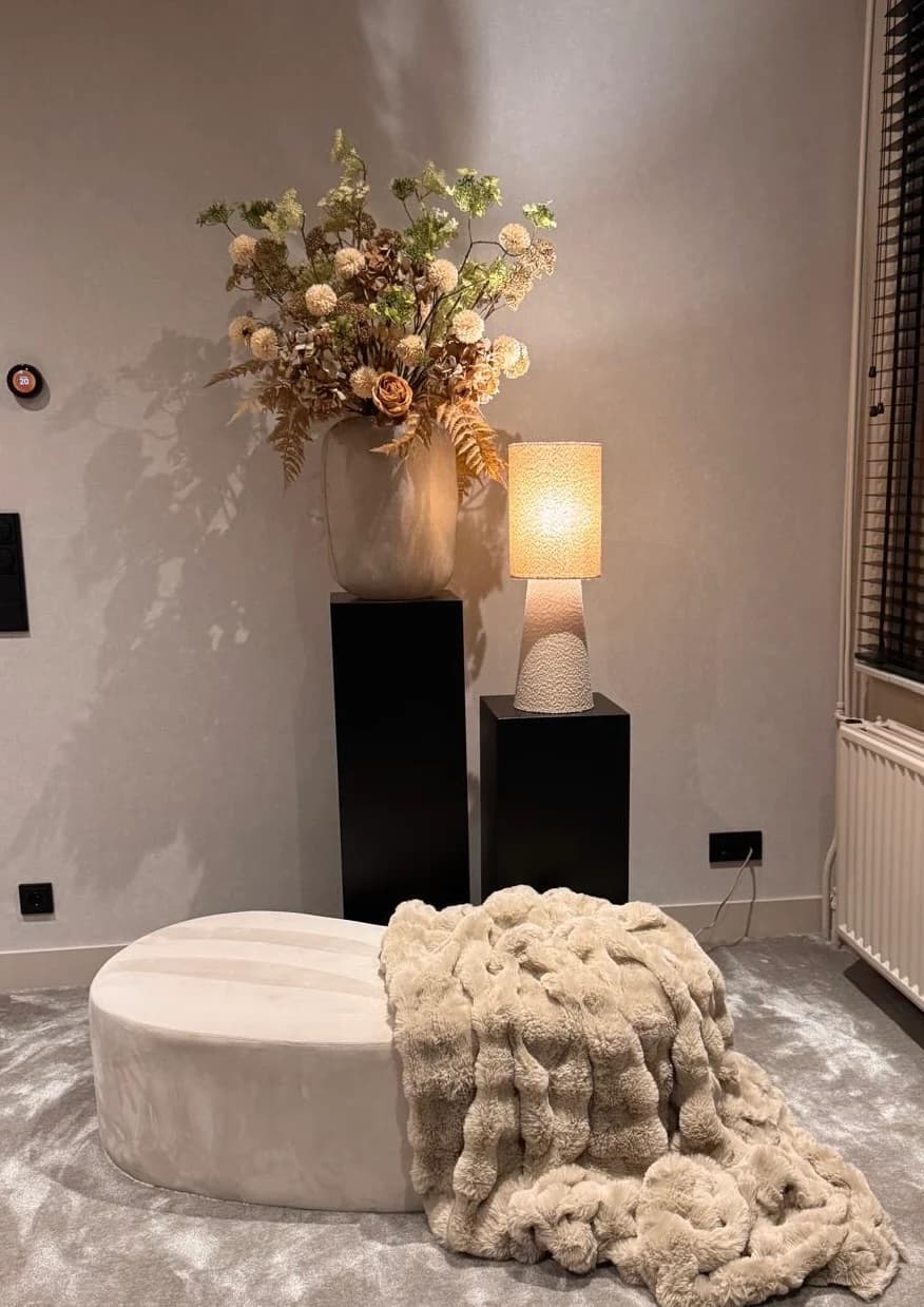 Droogboeket in zachte beige en roze tinten in een vaas op een zwarte zuil, naast een lamp in een woonkamerhoek