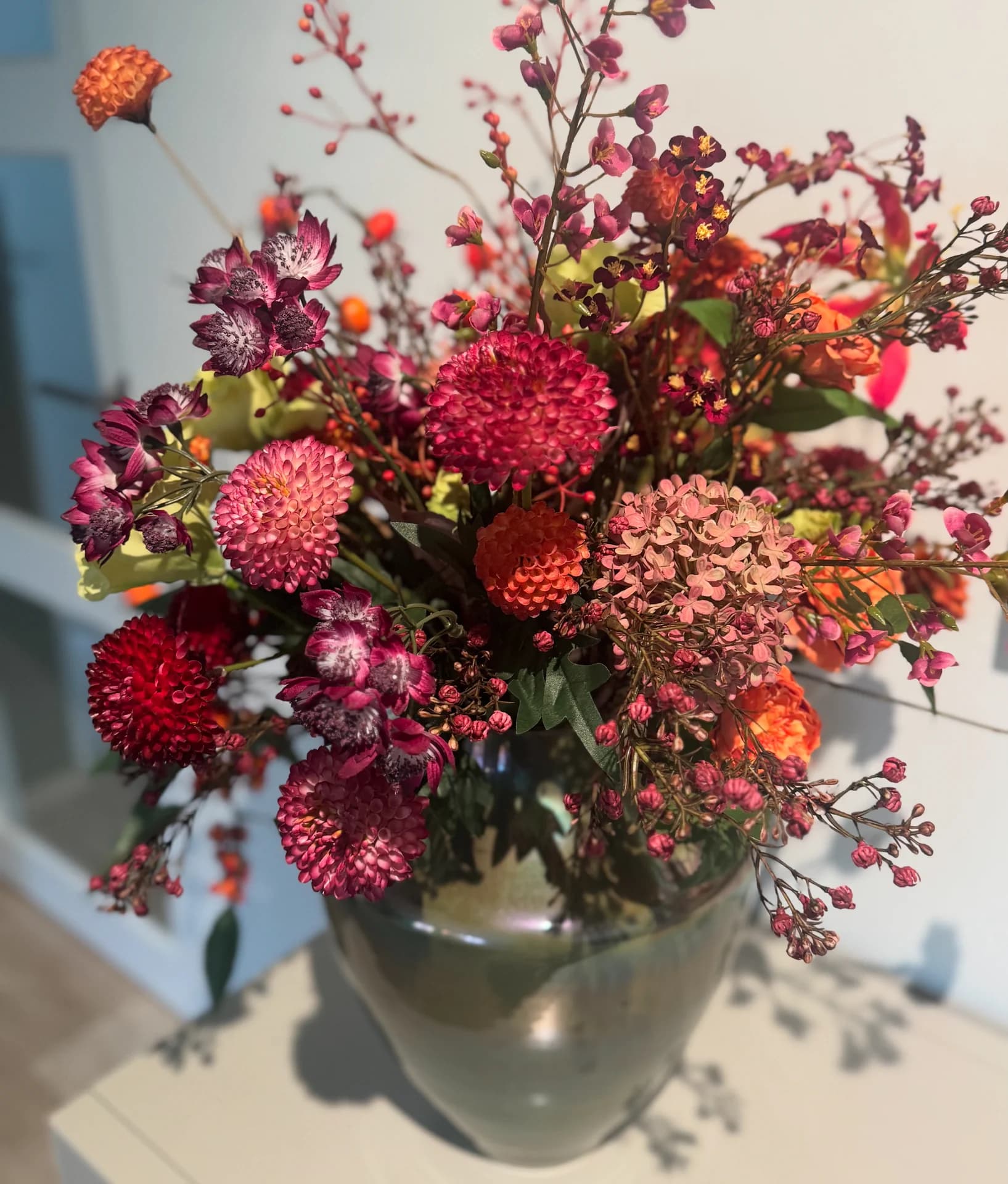 Compact boeket in rode, bordeaux en oranje tinten met dahlia's in een groenglazen vaas op een lichte tafel