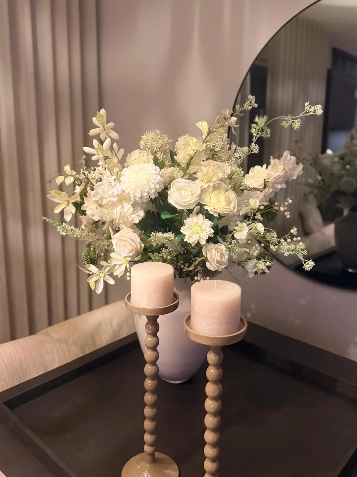 Wit zijden boeket met rozen en hortensia, tussen twee kaarsen op houten kandelaars voor een ronde spiegel
