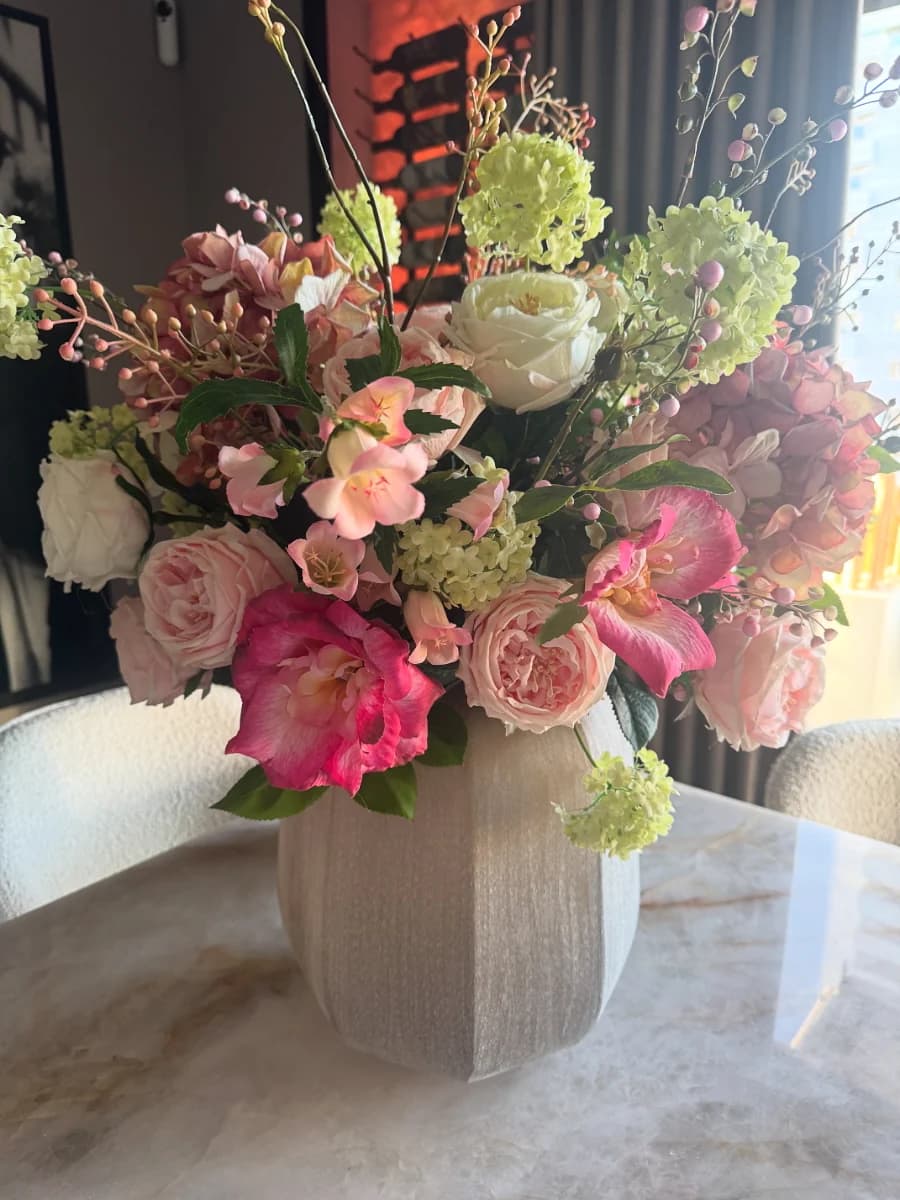 Ruim boeket met roze en fuchsia rozen, lichtgroene viburnum en pioenen in een beige geribbelde vaas op een marmeren tafel