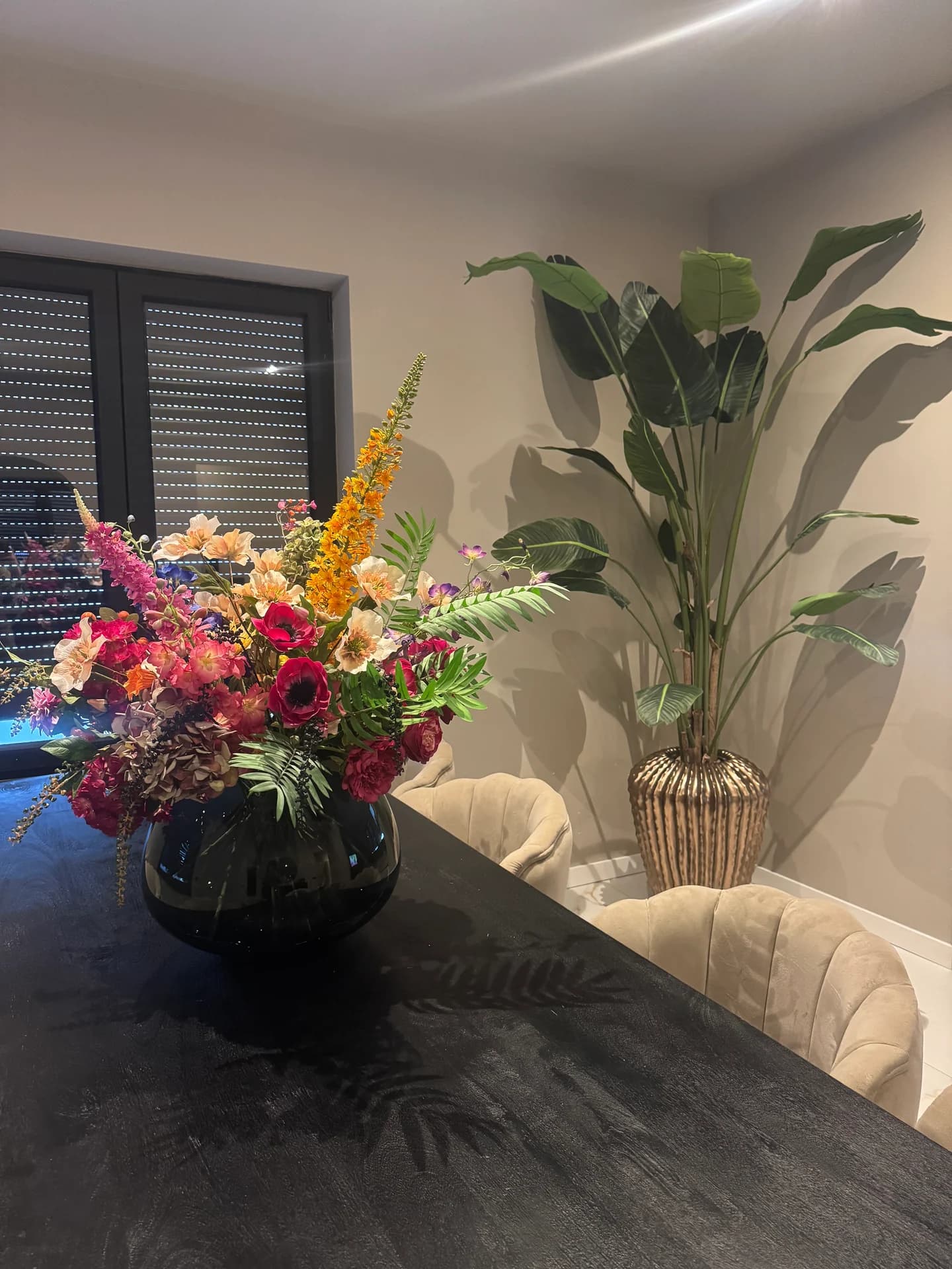 Boeket met oranje, roze en rode bloemen in een lage zwarte vaas op een donkere tafel naast een grote kamerplant