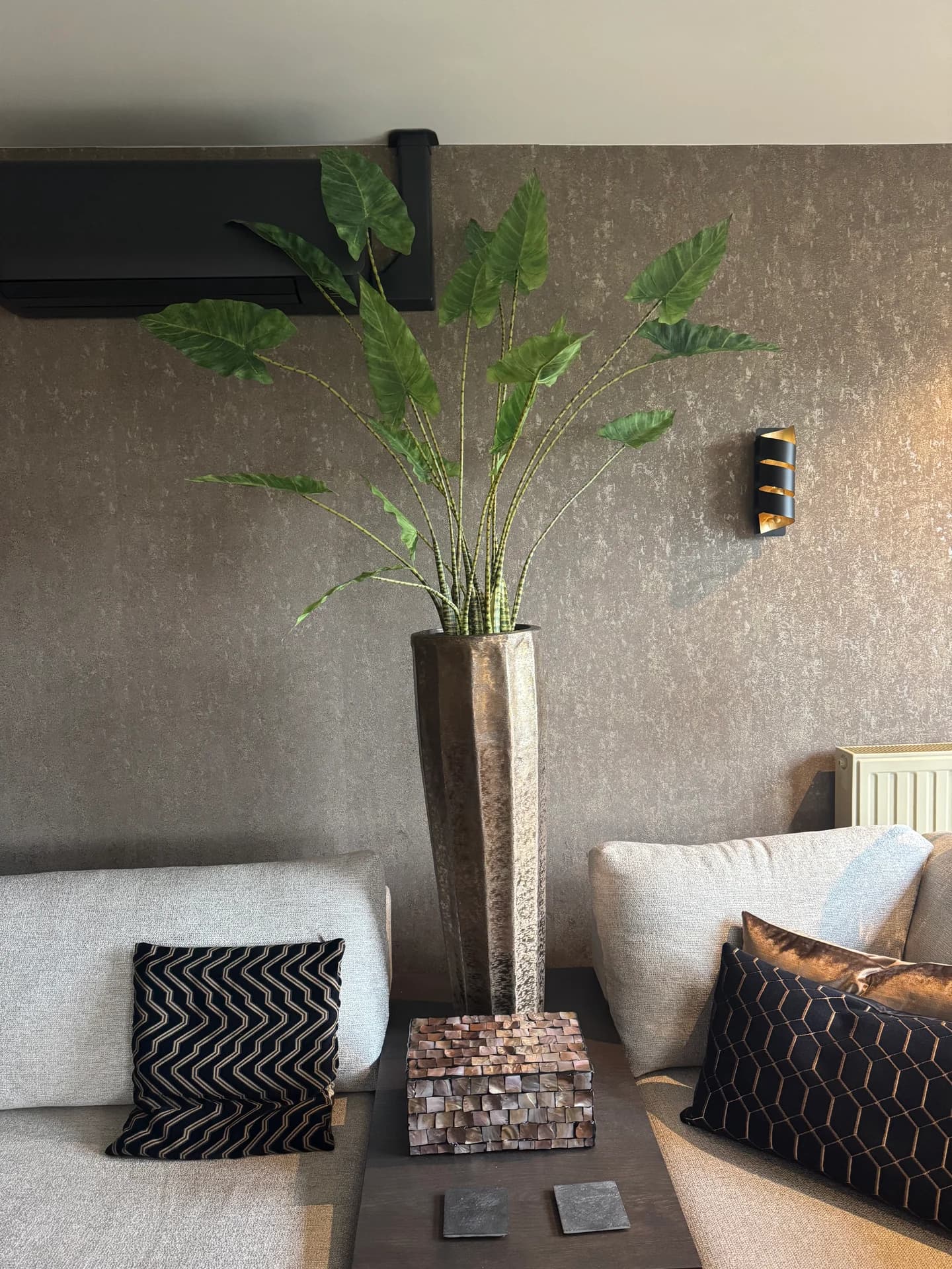 Grote groene bladplant in een hoge, smalle metalen pot tussen witte kussens op een bank, voor een bruine wand
