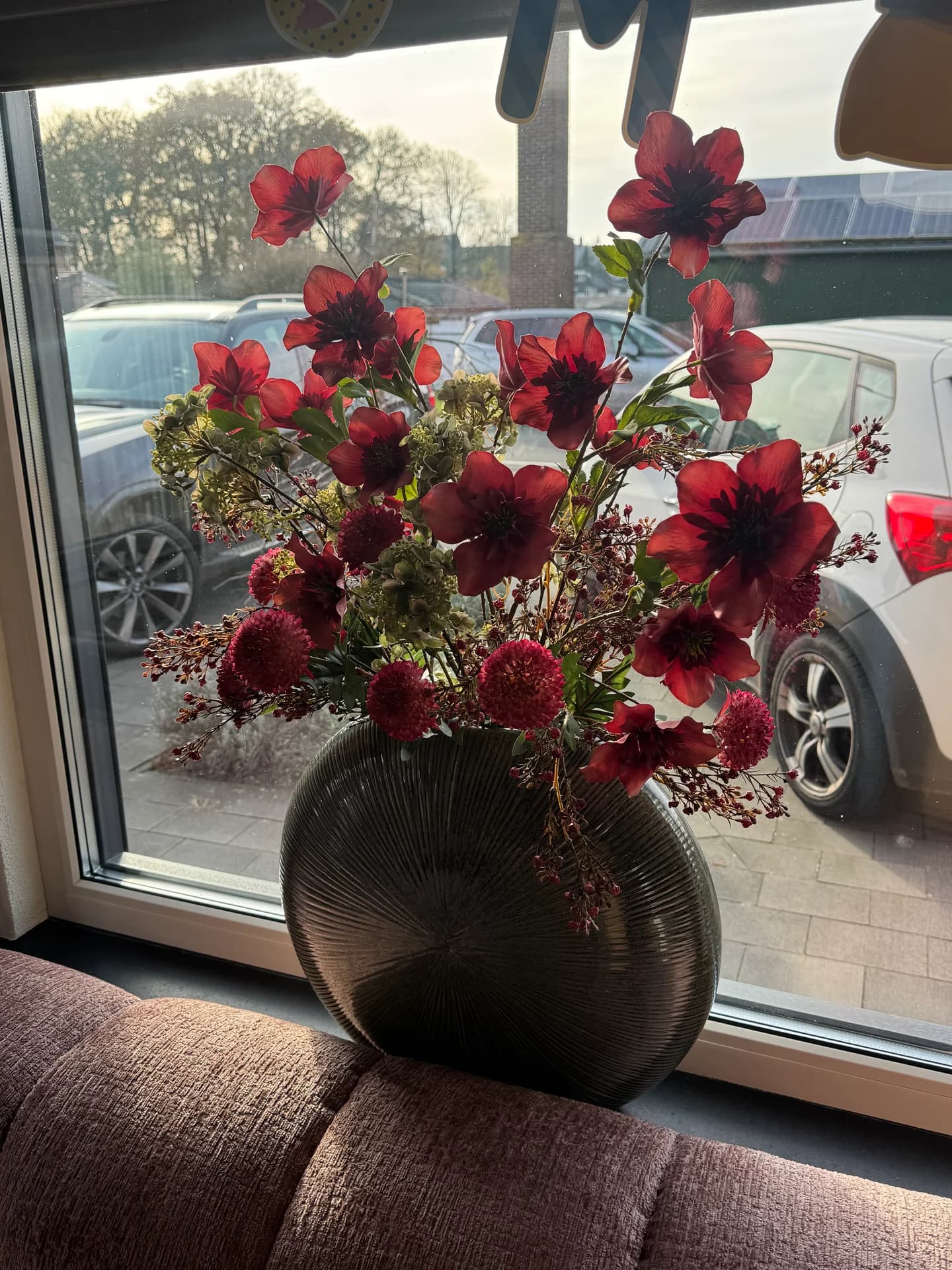 Donkerrood boeket met bordeaux bloemen in een ronde donkere vaas met ribbels op een vensterbank voor een groot raam