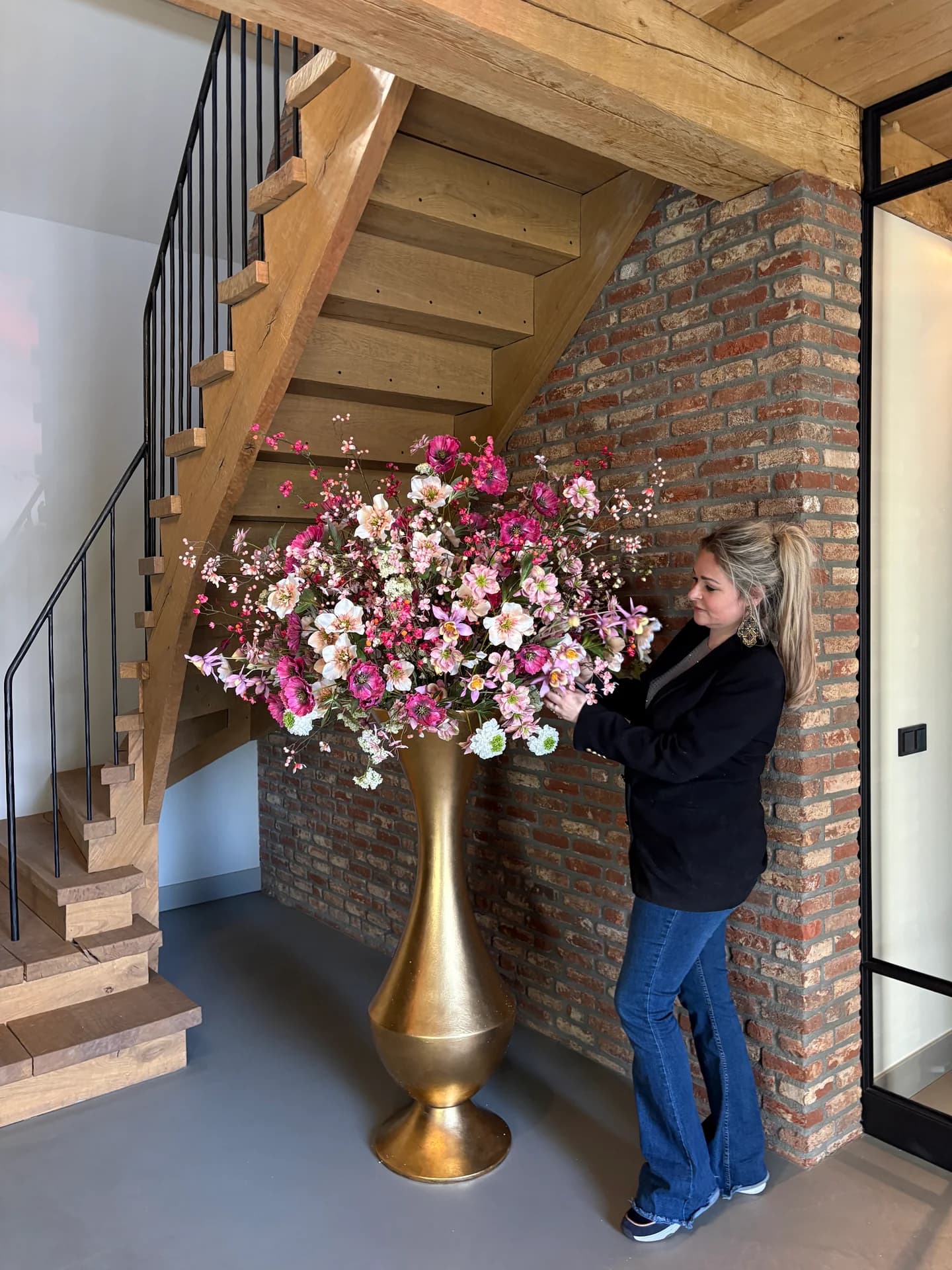 Een vrouw plaatst een roze-wit boeket in een hoge gouden vaas naast een houten trap en bakstenen muur
