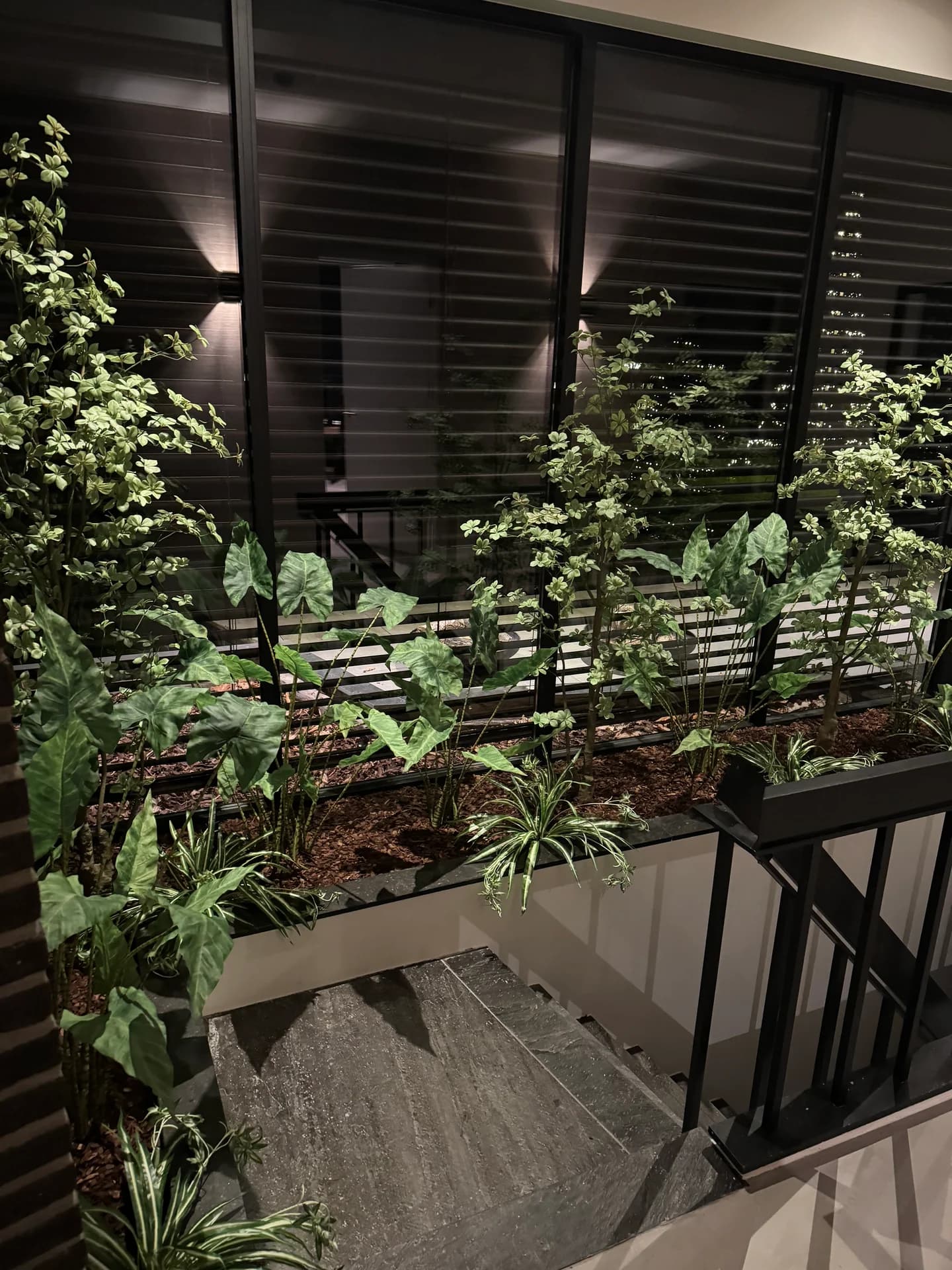 Groene kunstplanten en hangende bladeren in een plantenbak bij een zwarte trapleuning en zwarte jaloezieën