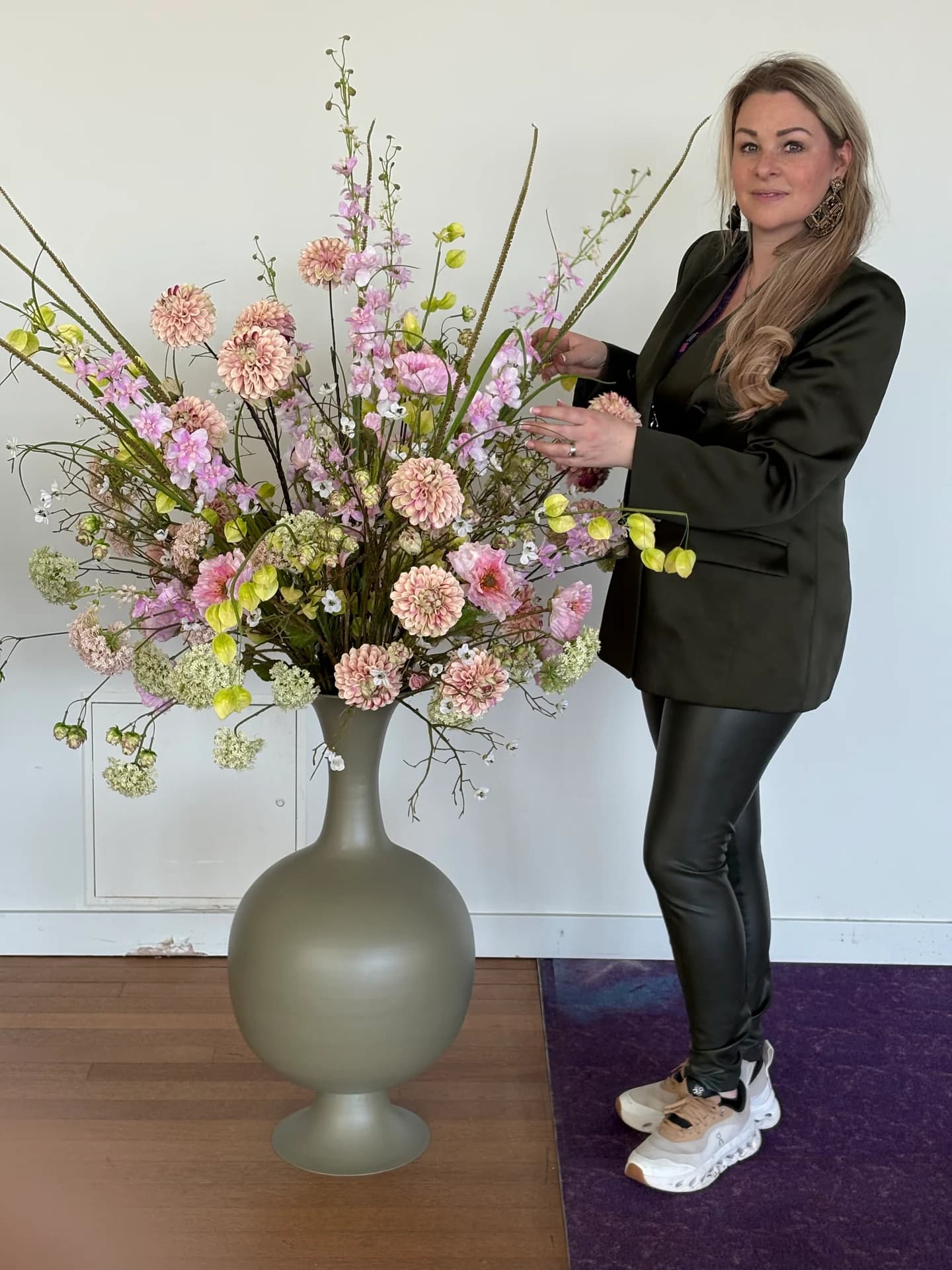 Een vrouw staat naast een hoge olijfgroene vaas met roze en lichtpaarse zijden bloemen op een houten vloer