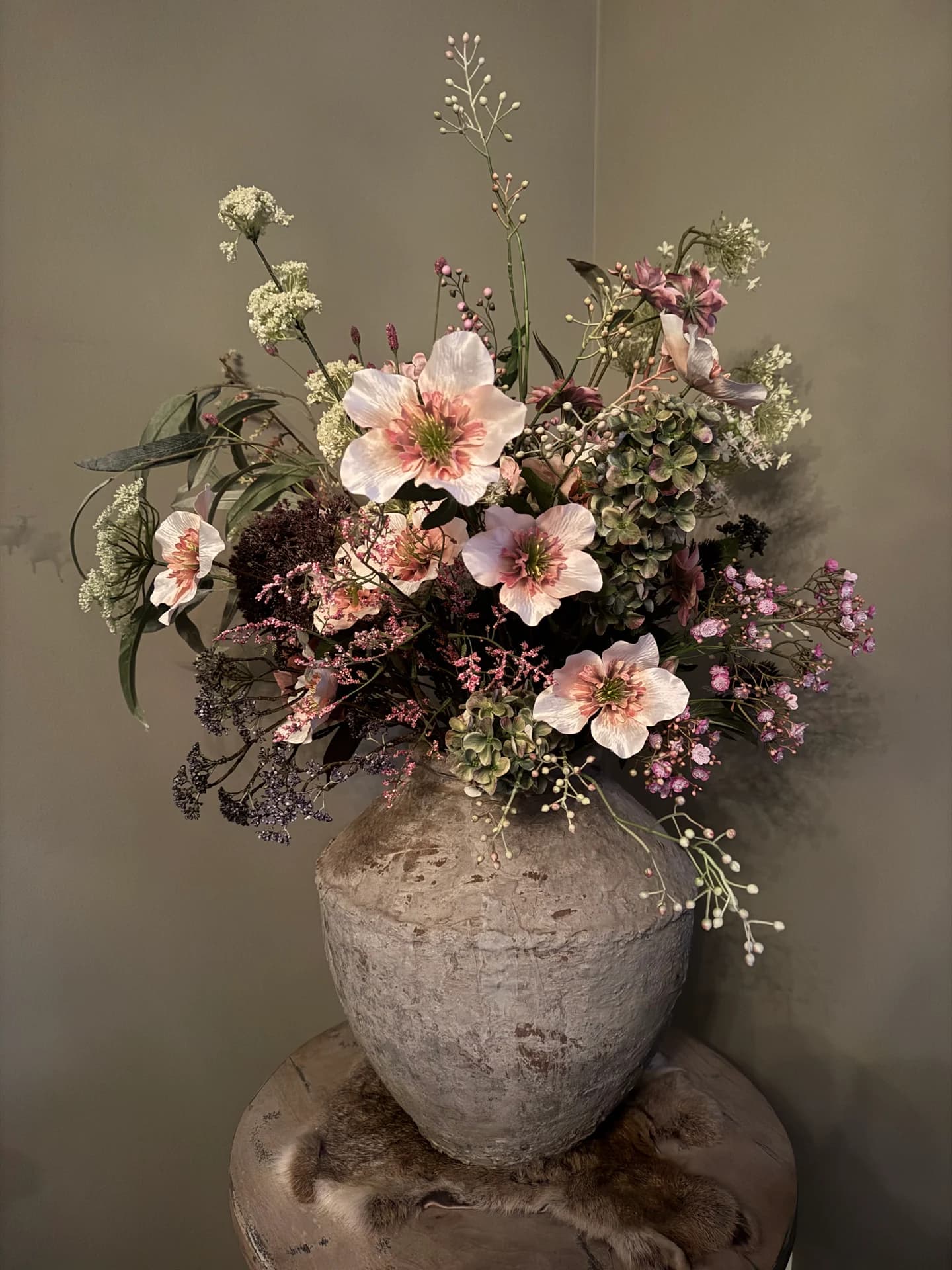 Boeket met roze bloemen, bruine bladeren en kleine bloemetjes in een grove stenen vaas op een houten voet